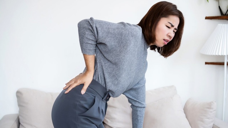 ソファから立ち上がる際に腰を押さえて痛みを訴える女性。ぎっくり腰の応急処置や翌日ケアの説明