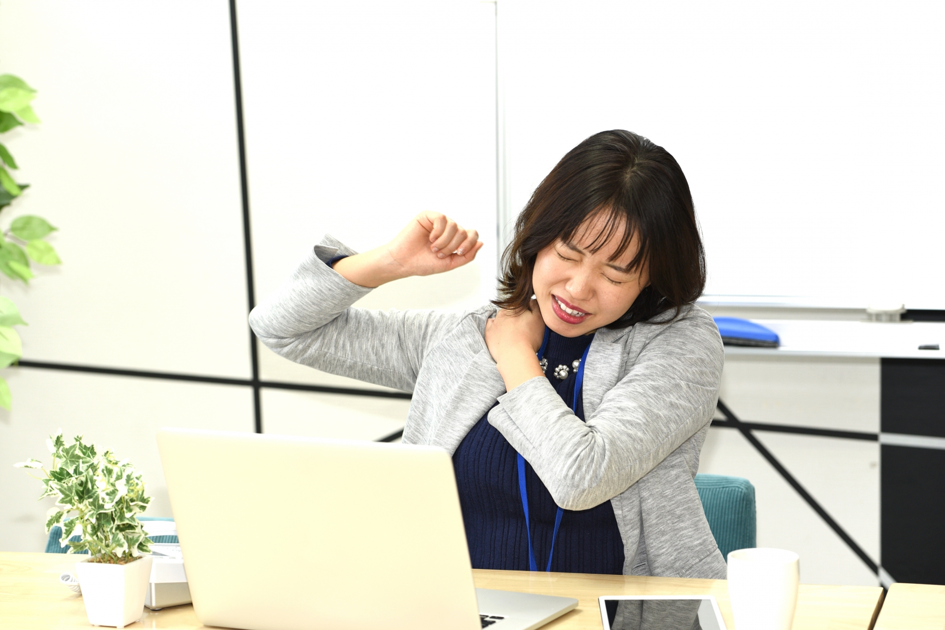 ノートPCの前で肩を押さえて痛そうにするオフィス女性。長時間座位で肩や背中がこわばっている様子