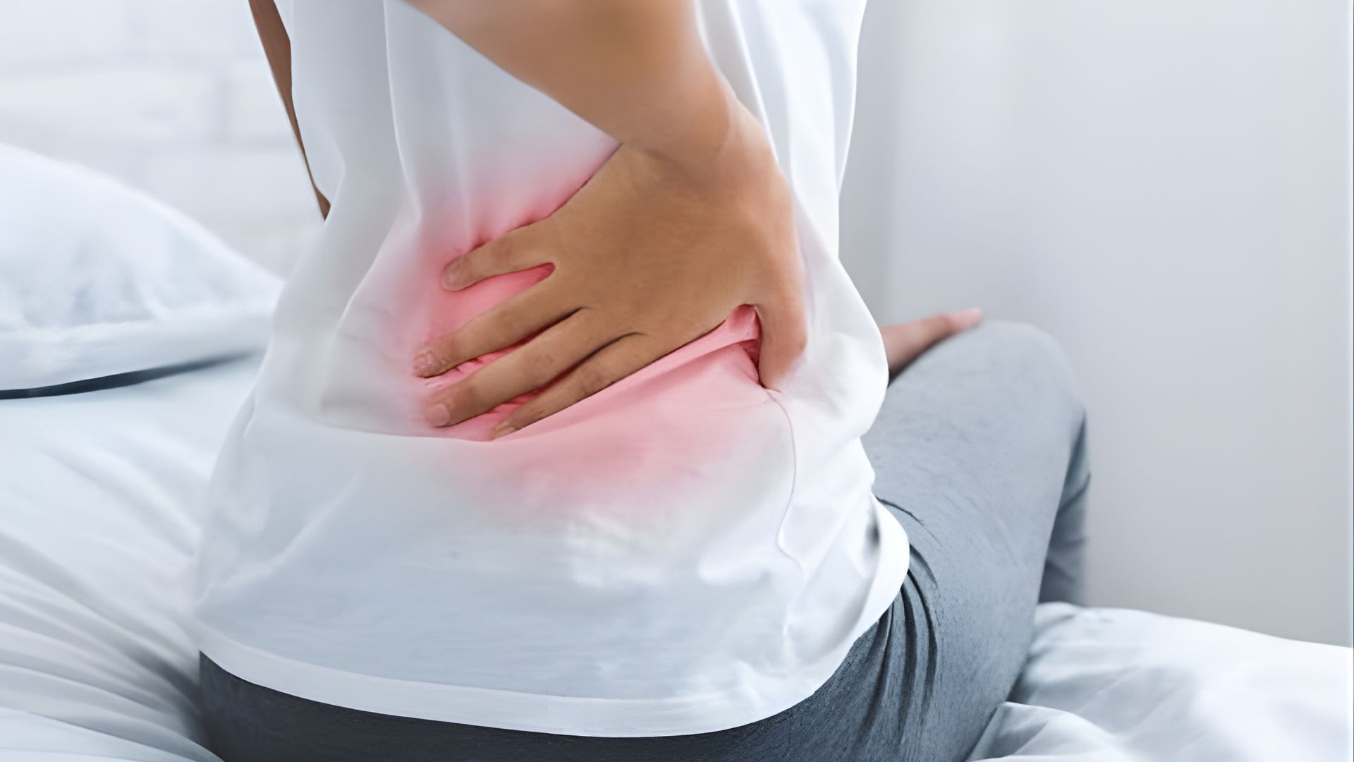 ベッドの端に座り、片手で腰を押さえる人物。腰の痛みが強く出ている様子